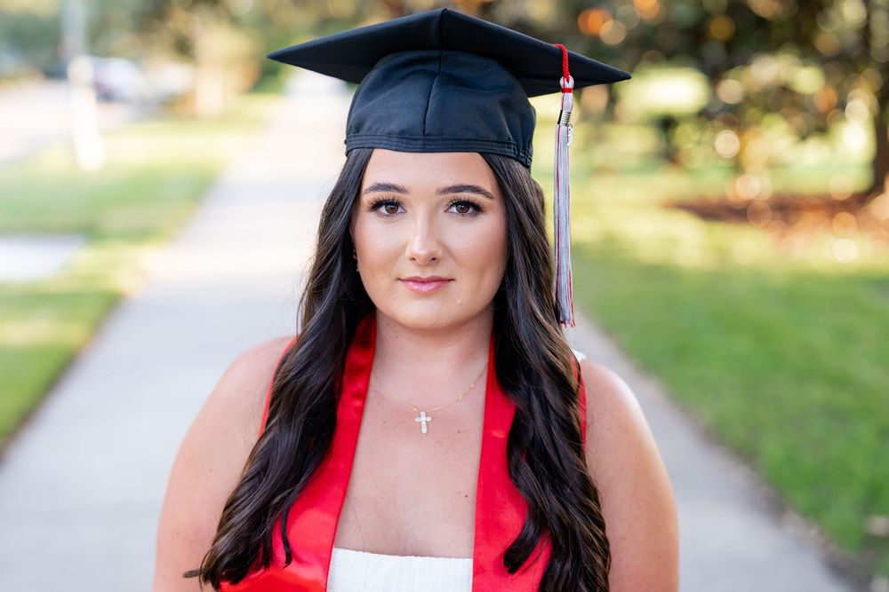 Young woman with long dark hair wearing a black graduation cap with tassel and red stole poses outdoors on a tree-lined path.