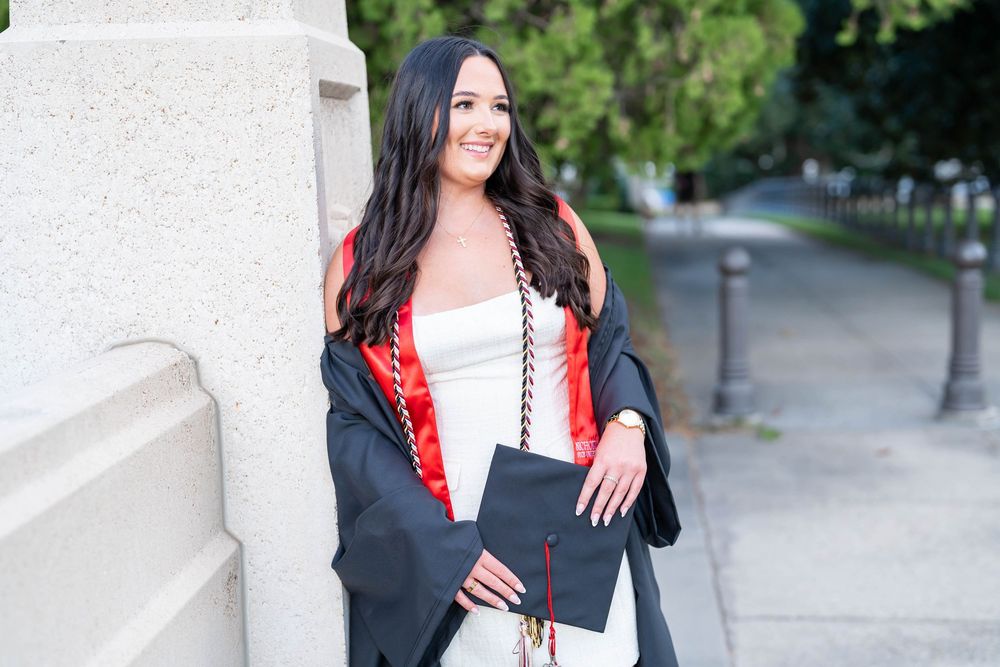 Smiling graduate in a white dress, black gown and red stole holding her cap and leaning against a stone pillar on a tree-lined path.
