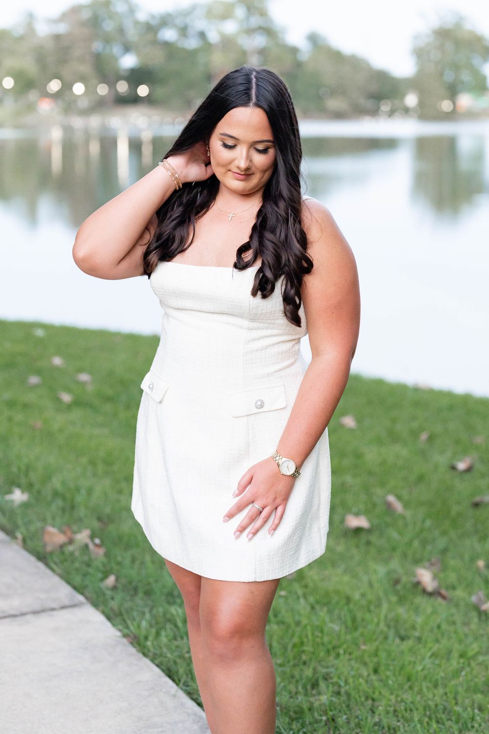 Woman with long dark wavy hair in a white strapless mini dress poses by a calm lake on a grassy lawn scattered with fallen leaves.