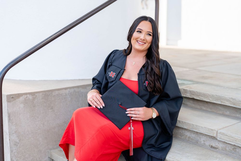 Smiling graduate in a black gown and red dress sits on outdoor stone steps holding a black graduation cap with a red tassel.
