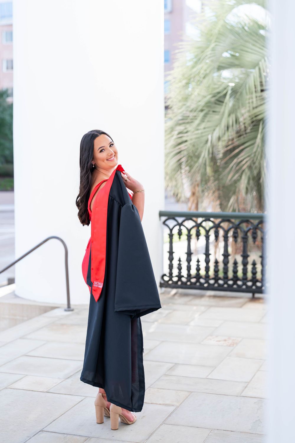 Smiling graduate in a black gown with a red stole, holding her cap and gown over her shoulder beside a palm tree and white columns.