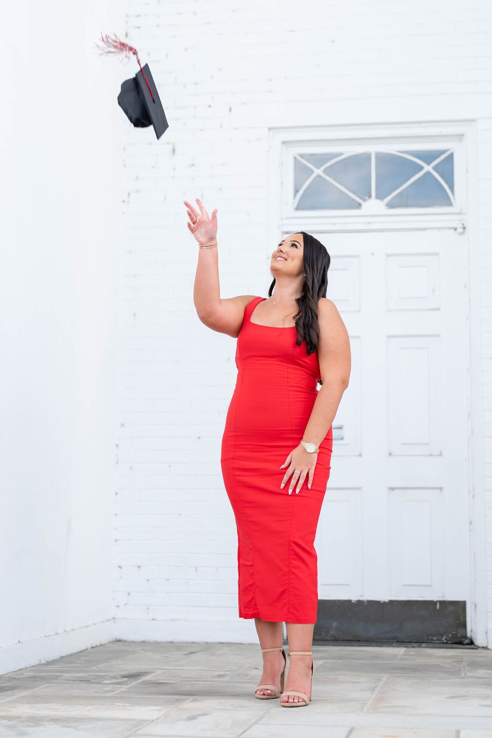 Woman in a red dress and heeled sandals tosses her black graduation cap into the air in front of a white brick building.