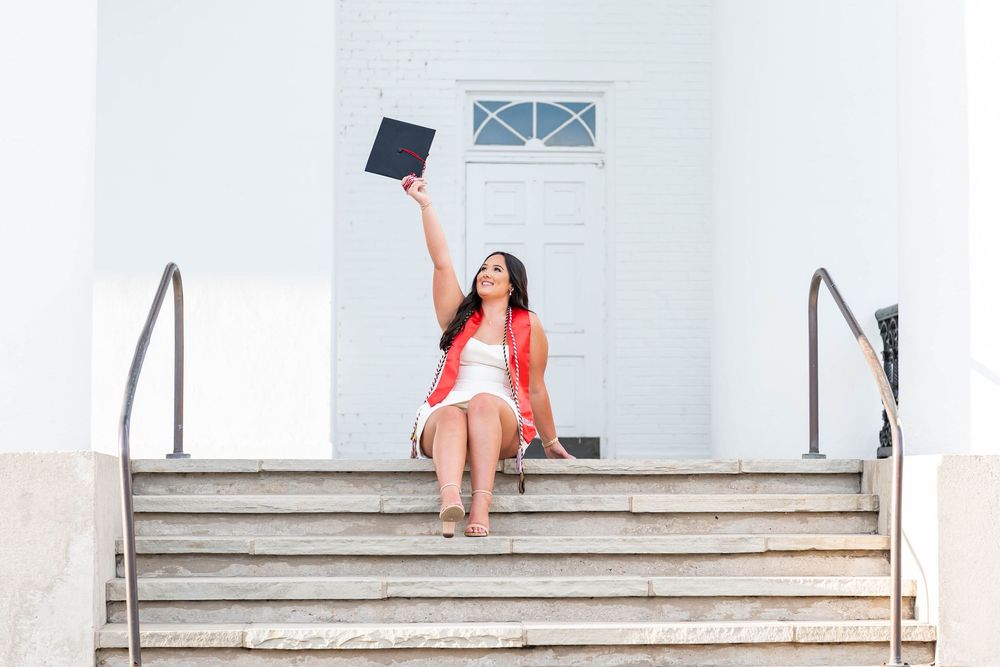 Smiling graduate in a white dress and red cardigan sits on white church steps holding a graduation cap high in the air.