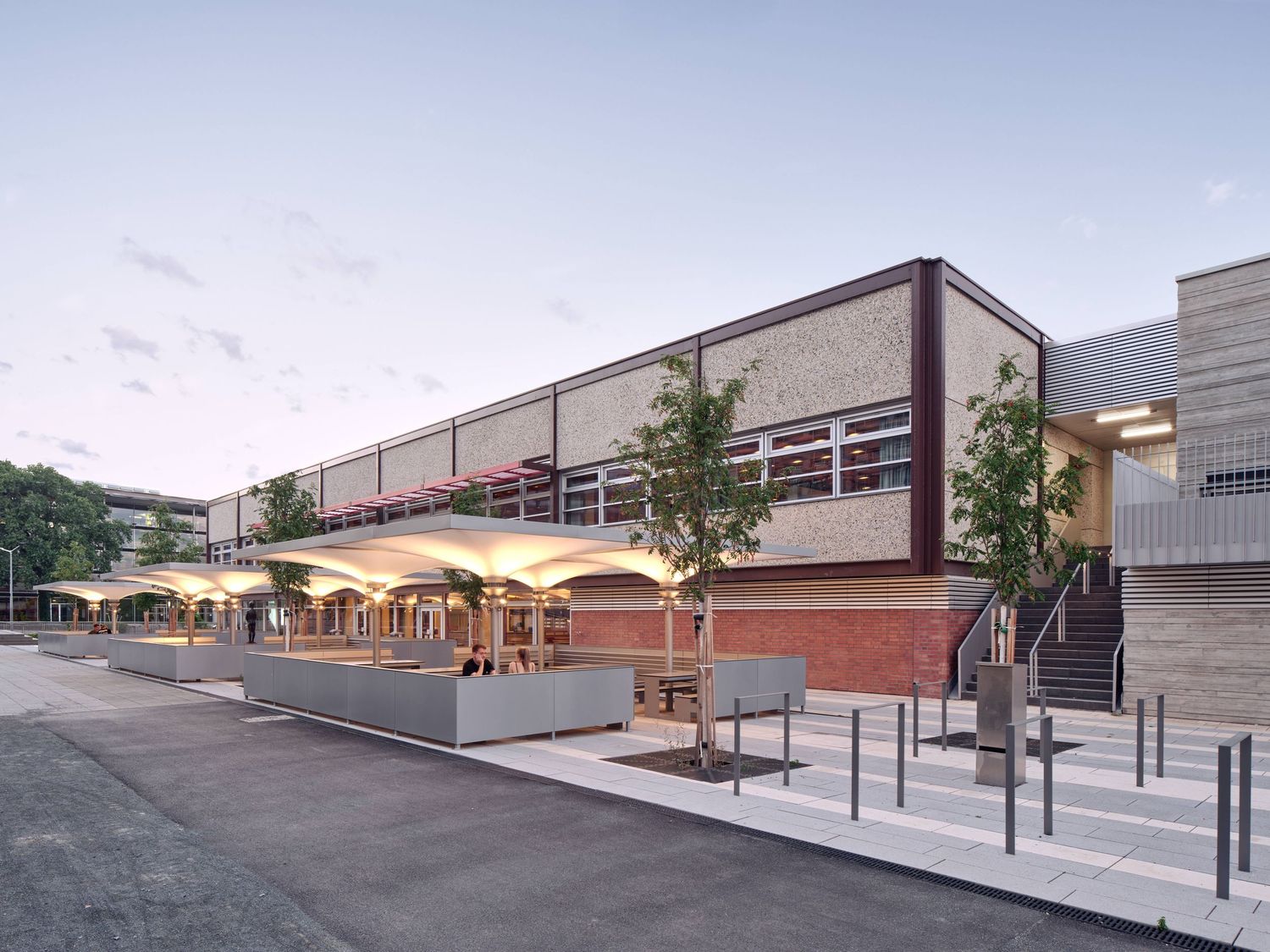 Modern brick and concrete market building at dusk with a white canopy, outdoor seating, young trees, and a paved plaza.