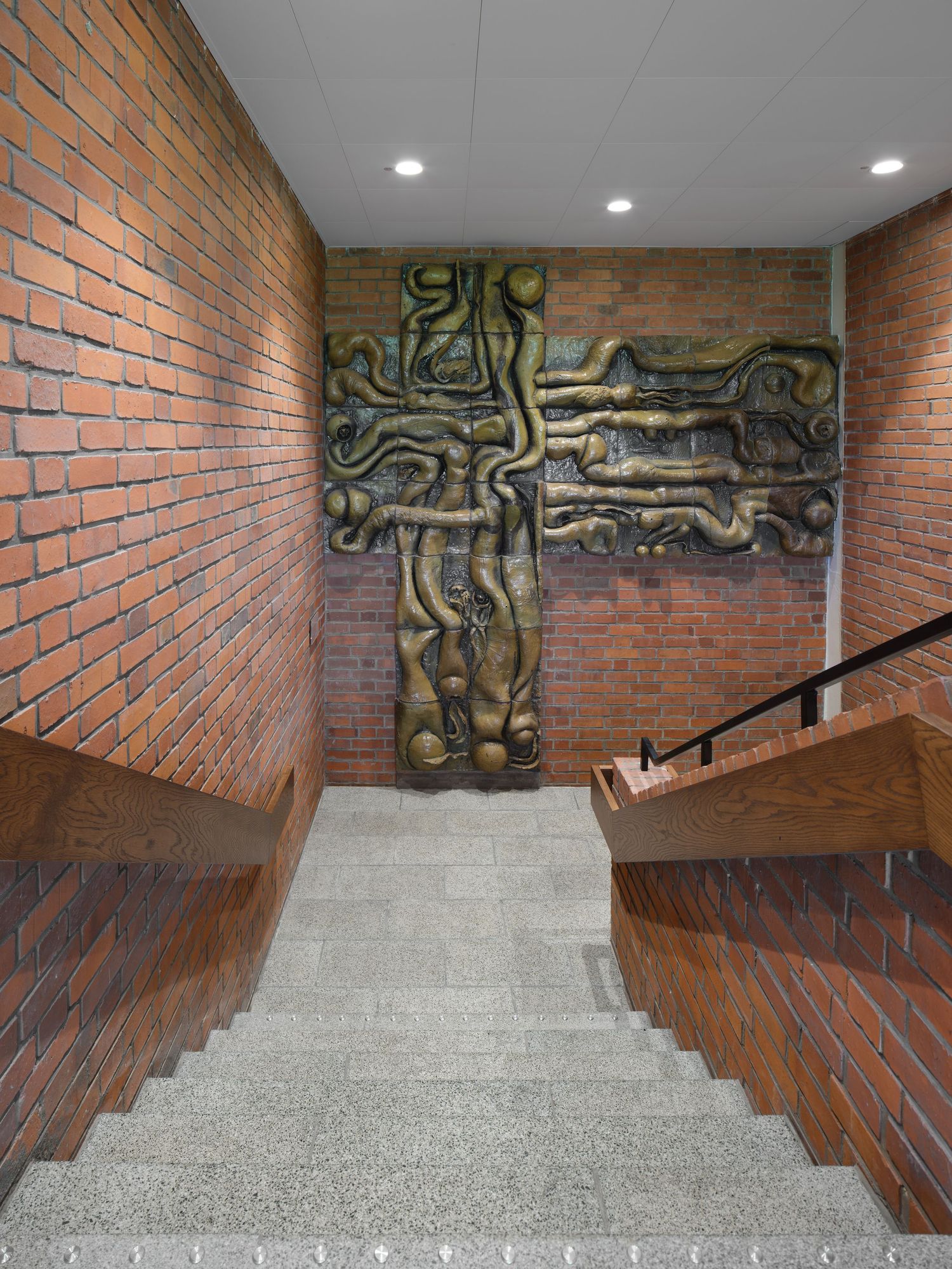 Red brick stairwell with granite steps and a wooden handrail leading down toward a large bronze cross-shaped relief sculpture on the wall.