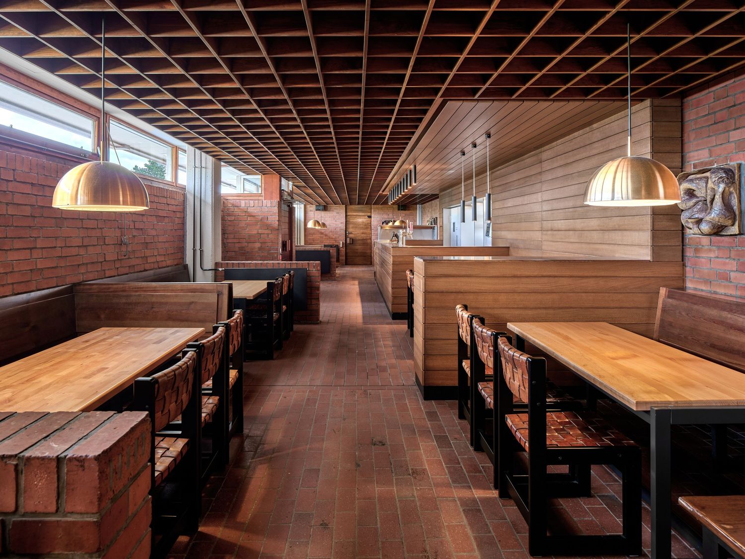 Warm modern restaurant interior with a wooden lattice ceiling, brick walls, pendant lamps, built-in booths and rows of rustic wood tables.