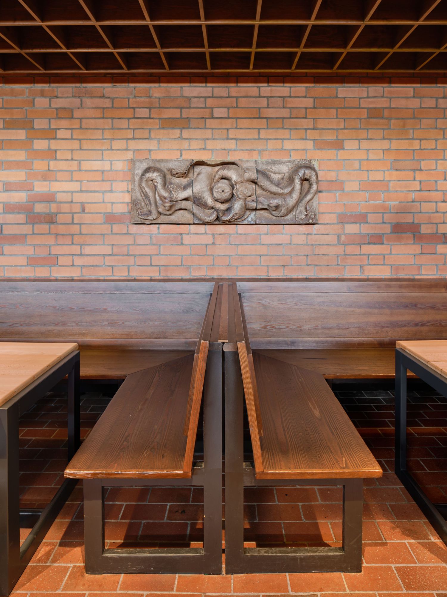 Rustic restaurant interior with dark wooden booth seating, a red brick wall, and a carved octopus relief above tables on a brick floor.