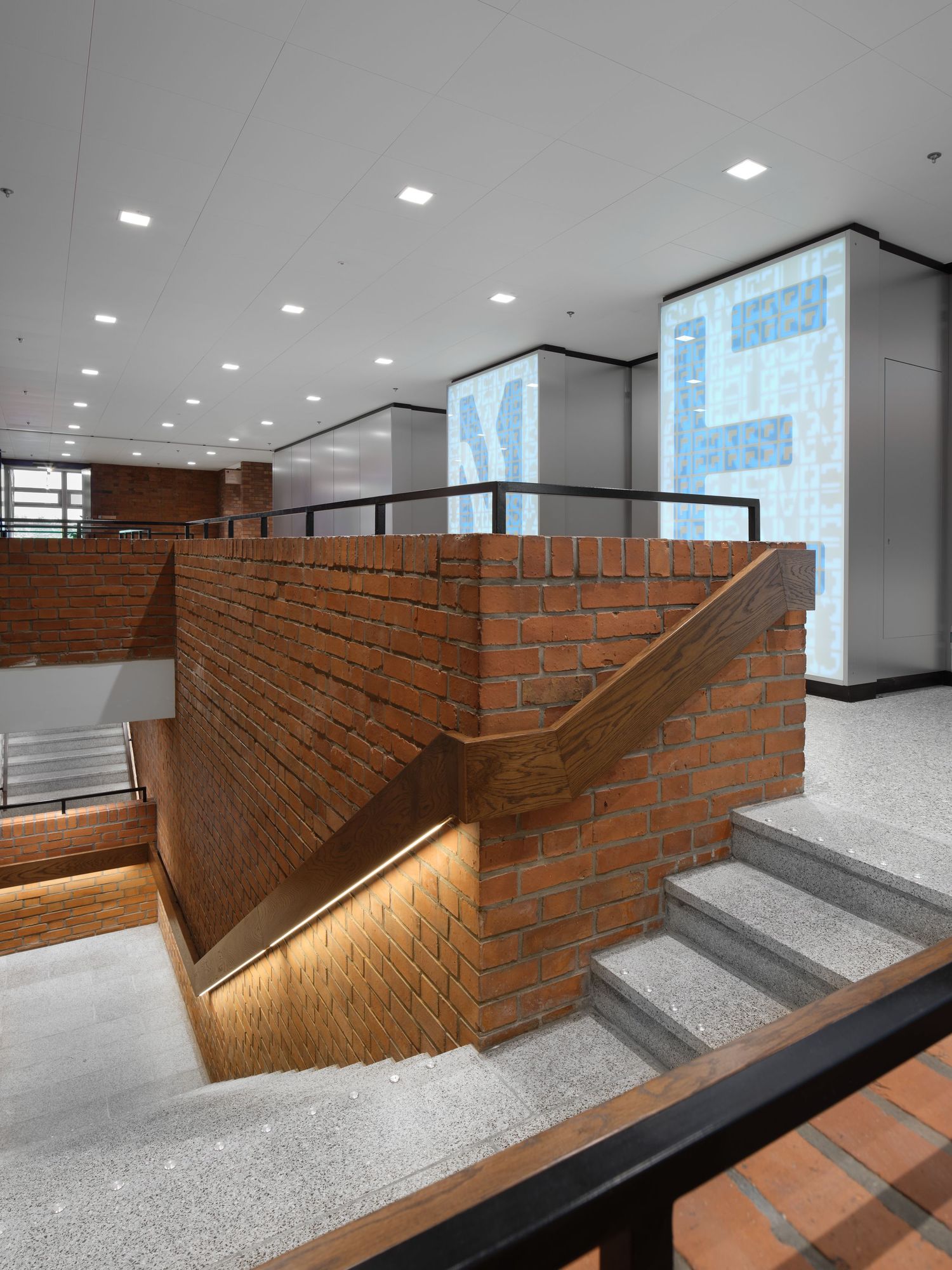 Modern interior staircase with red brick walls, a wooden handrail with warm lighting, speckled stone steps and illuminated glass panels.