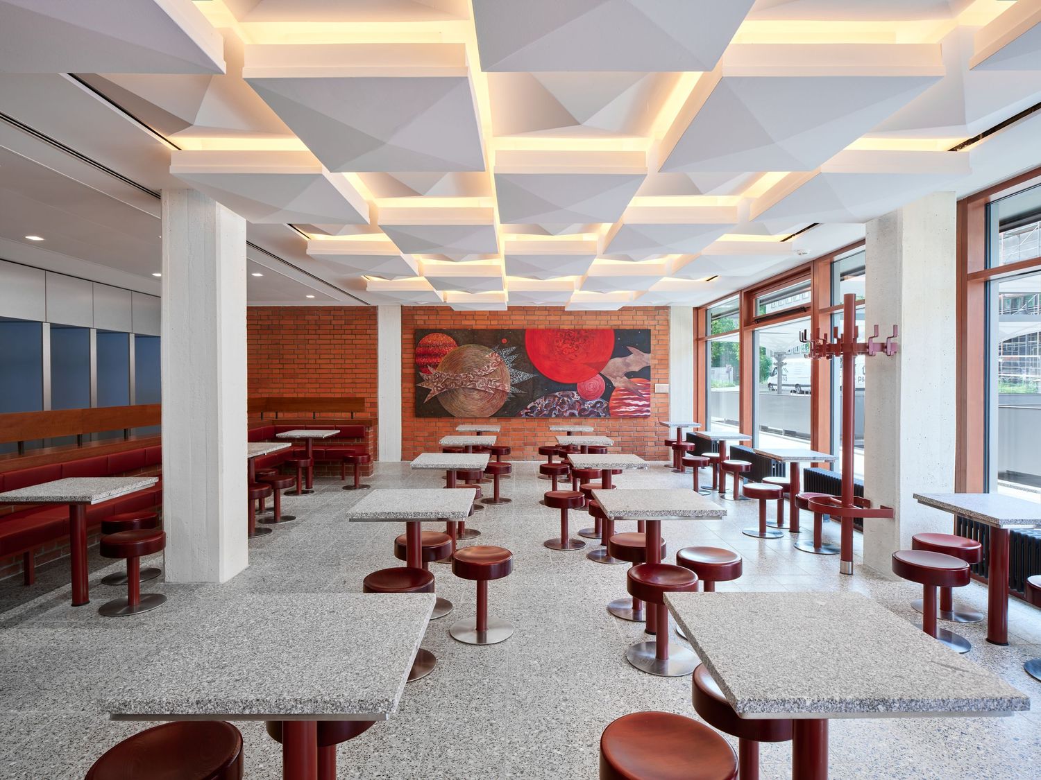 Modern restaurant interior with terrazzo tables, red stools, geometric white ceiling panels, red booth seating and a large colorful mural.