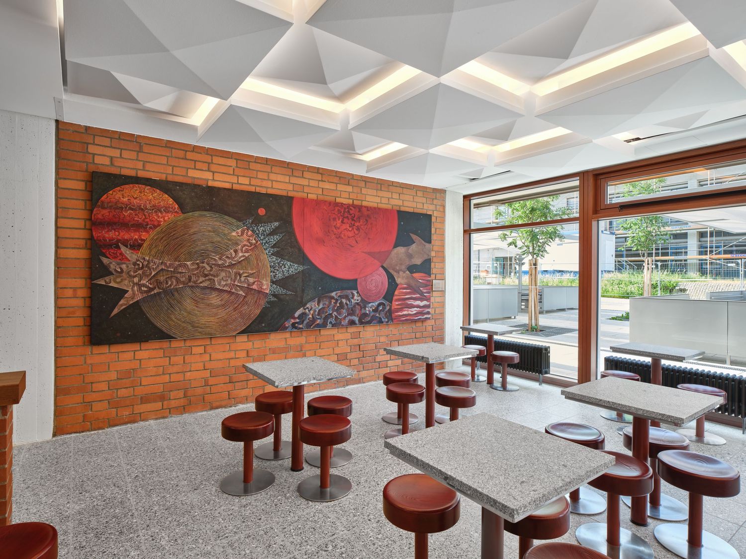 Modern cafe interior with red stools, speckled tables, a bold planetary mural on an exposed brick wall and a geometric white ceiling.