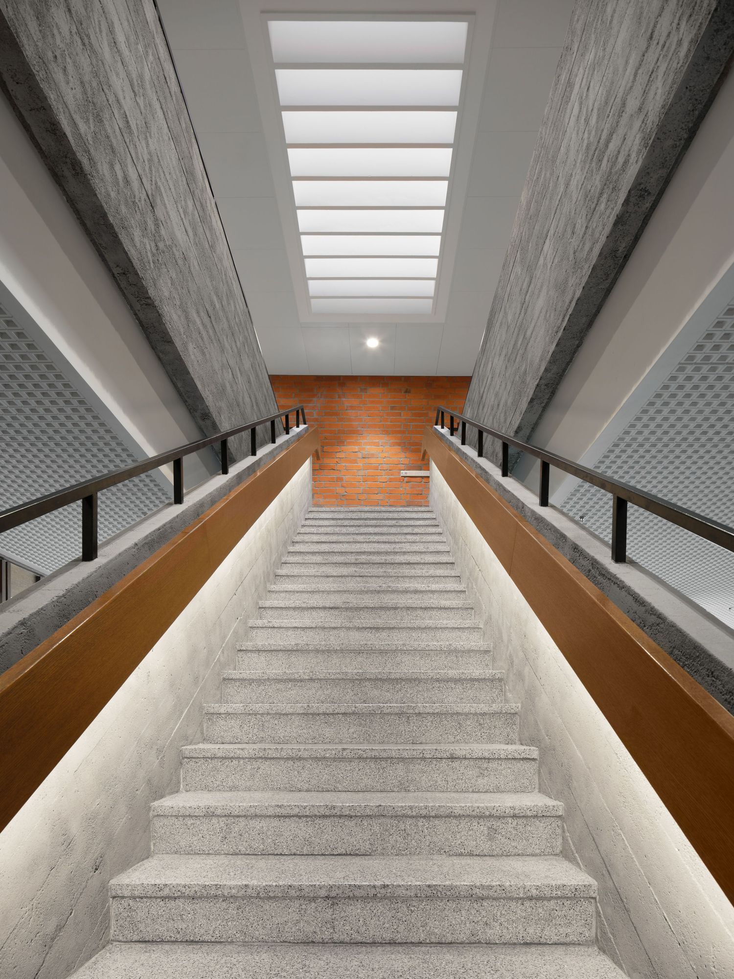 Symmetrical concrete staircase with wooden handrails leads up to a brick wall and a bright skylight in a modern architectural interior.