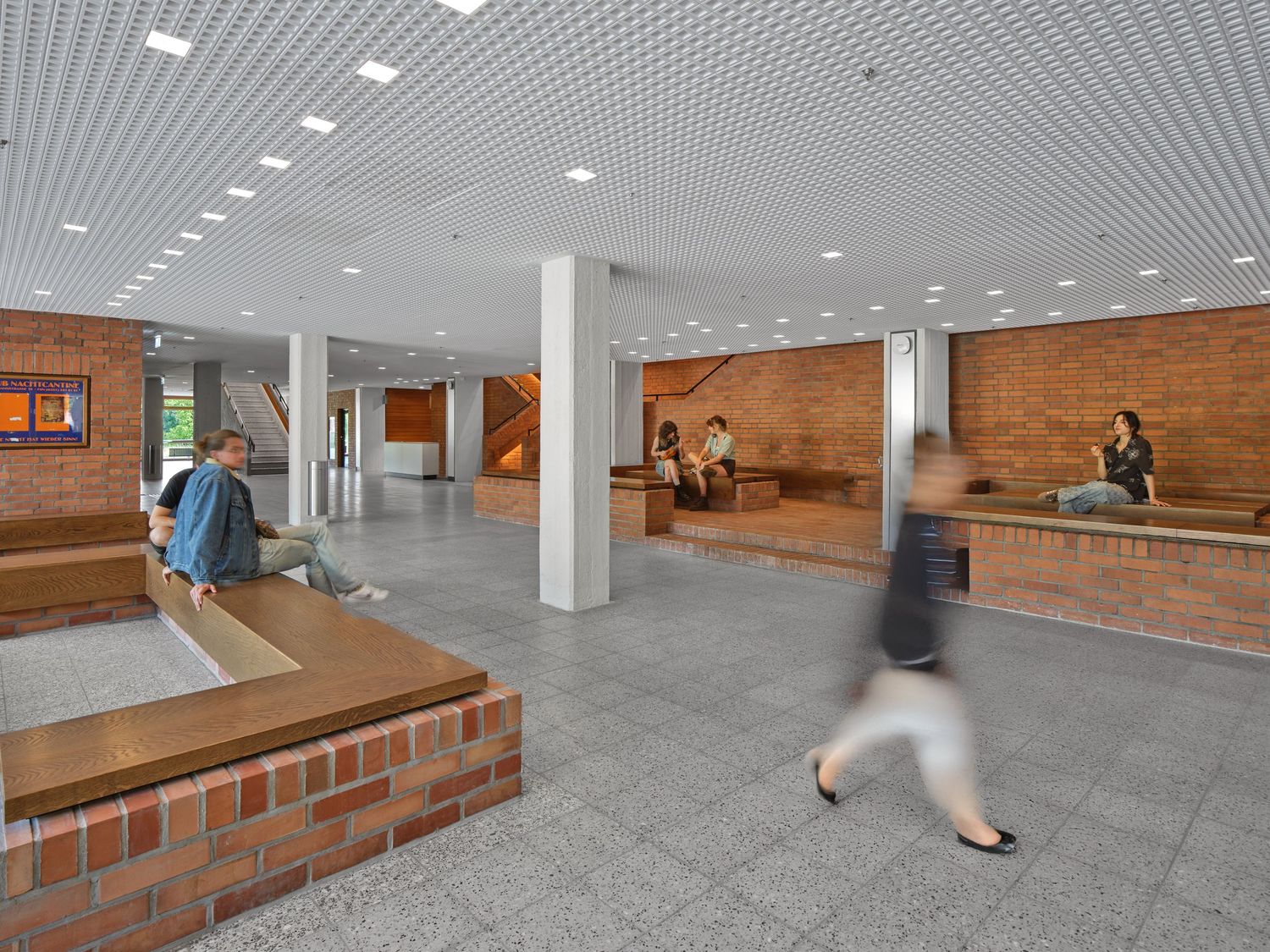 Spacious modern lobby with brick walls, white columns, and a perforated ceiling as students sit on built-in benches while a person walks by.