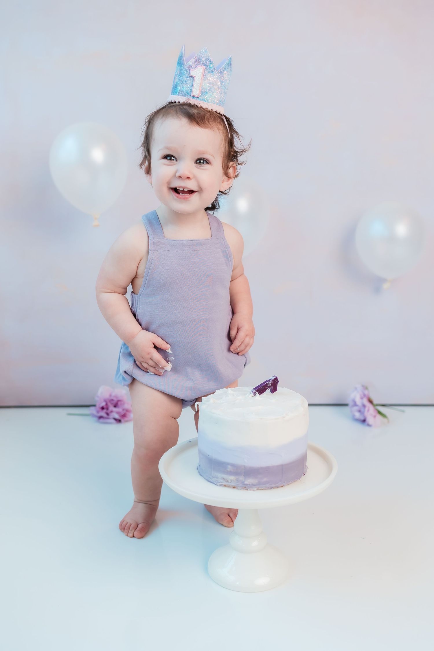Smiling baby in purple romper and crown celebrates first birthday with white cake and balloons.