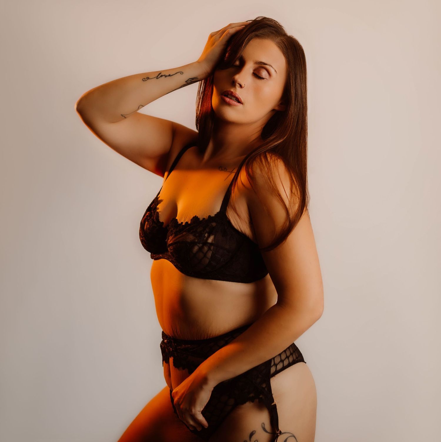 Woman in black lace lingerie set posing confidently against a light background with warm amber lighting.