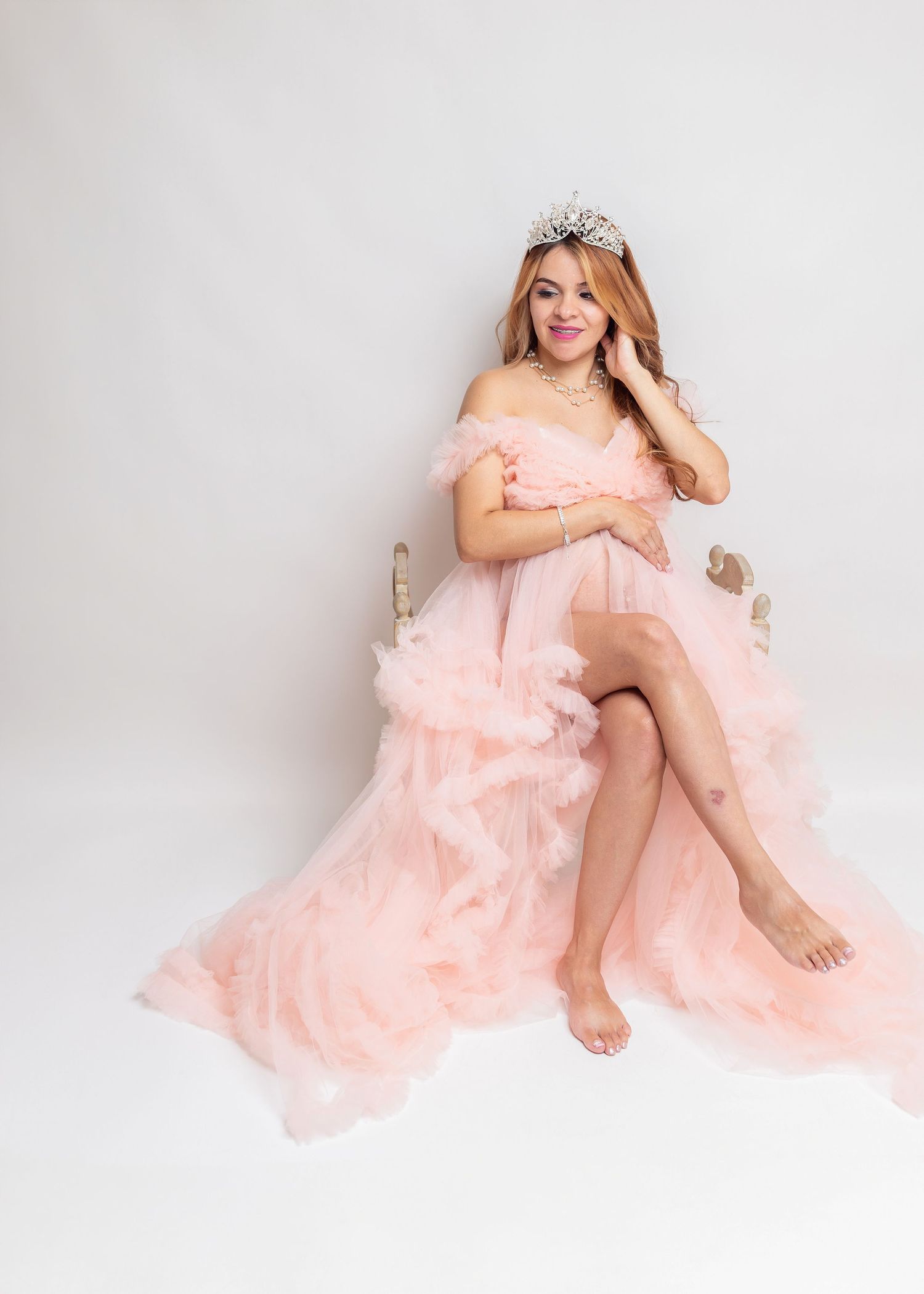Woman in pink tulle off-shoulder gown and crystal tiara poses gracefully on chair against white background.