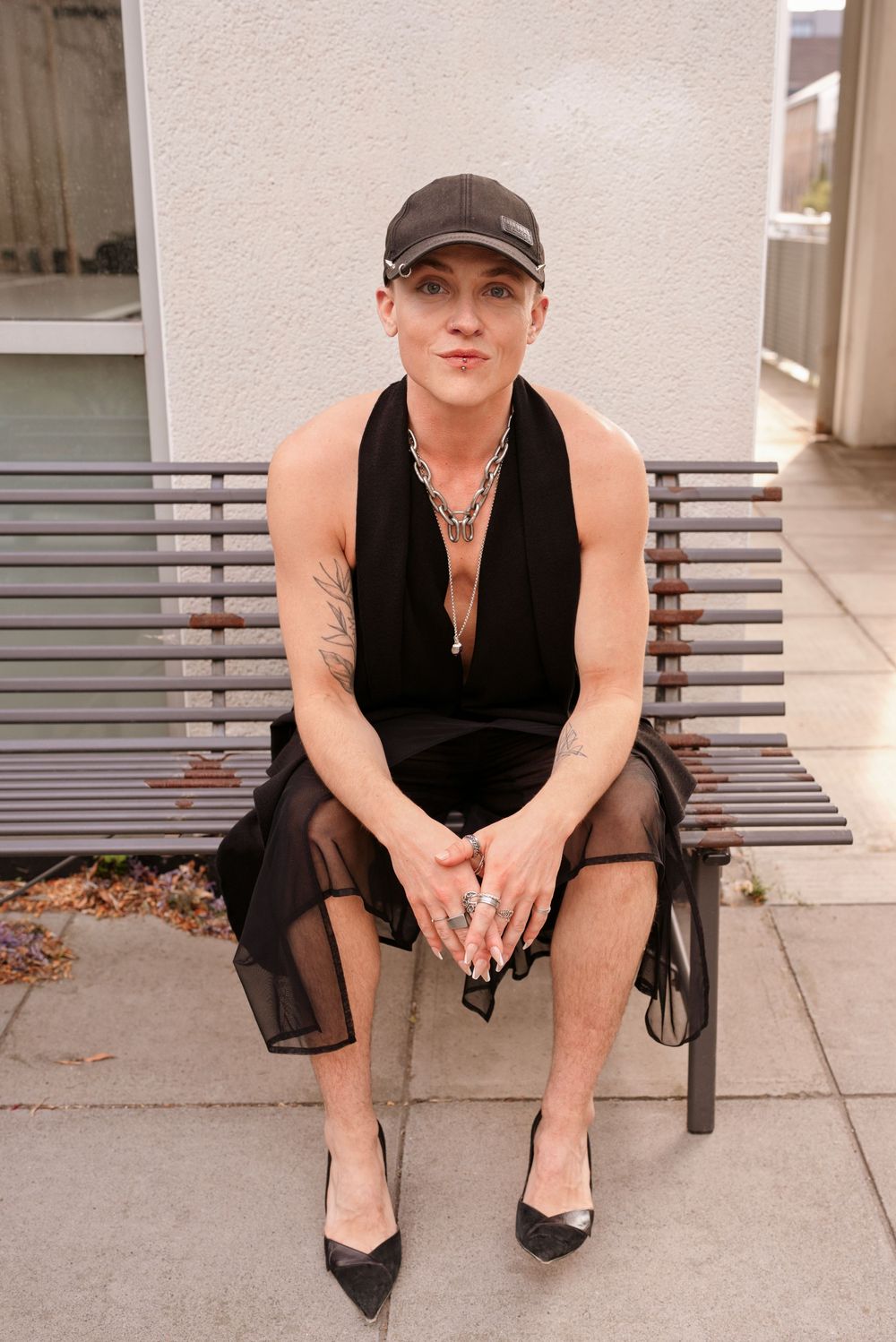 Person in black halter dress, cap, heels, and chain necklace sitting on bench outdoors.