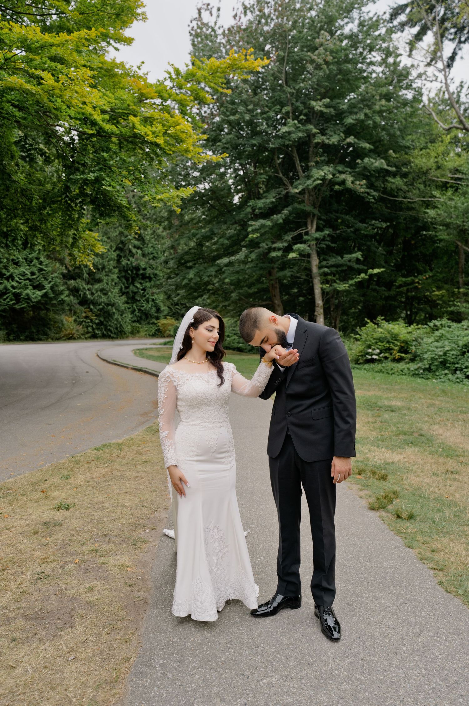 Groom in a black suit kisses the hand of his bride in a lace wedding gown and veil on a park path surrounded by green trees.
