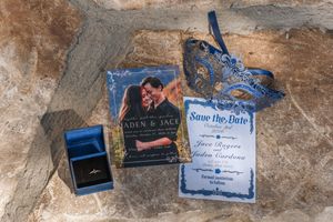 Wedding save-the-date cards, engagement ring in blue box, and decorative masquerade mask displayed on stone surface.