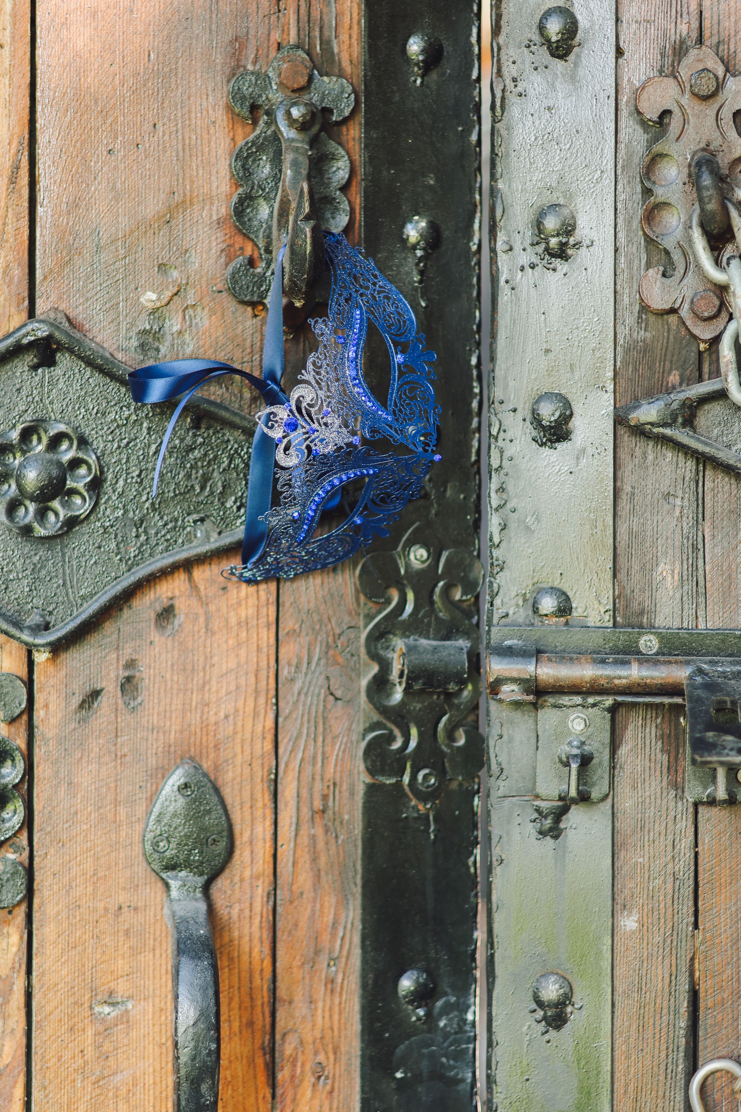 Blue ornate Venetian masquerade mask hanging on an antique wooden door with vintage iron hardware and locks.
