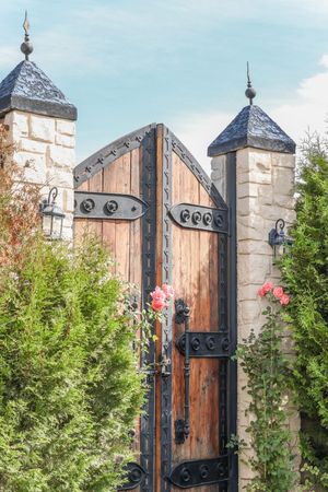 Ornate wooden garden gate with black wrought iron details flanked by stone pillars and pink roses.