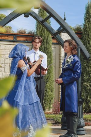Couple exchanges vows outdoors in blue wedding attire under arch with officiant at garden ceremony.