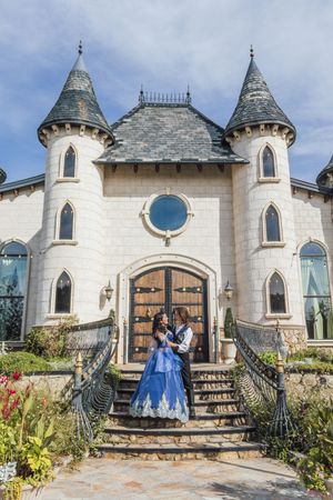 Couple in royal costumes posing on castle steps with white turrets and ornate wooden doors under blue sky.
