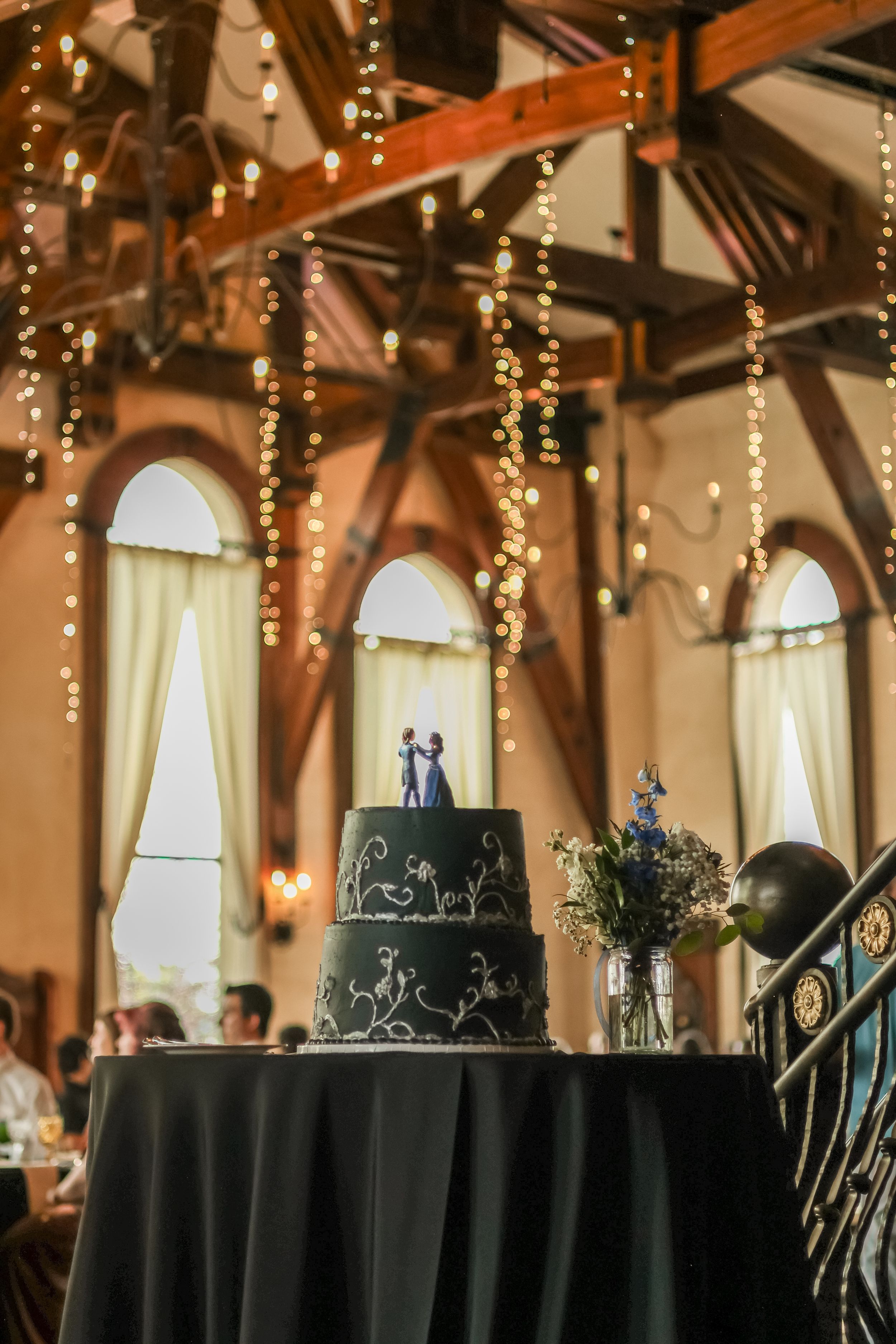 Elegant three-tier black wedding cake with floral design and couple topper displayed in festive venue.