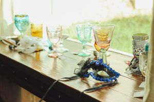 Colorful vintage glassware and a blue masquerade mask displayed on a wooden table near a bright window.
