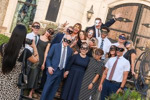 Large group of people wearing masquerade masks posing together outdoors at a formal event near an elegant building.
