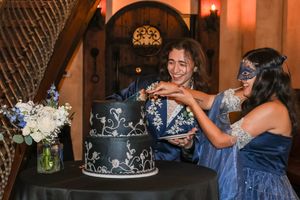 Two teens in blue masquerade attire cut a elegant black tiered cake with white floral designs at a formal celebration.