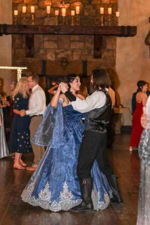 Couple dancing in medieval-style attire at a wedding reception, woman in blue lace gown, stone fireplace backdrop.