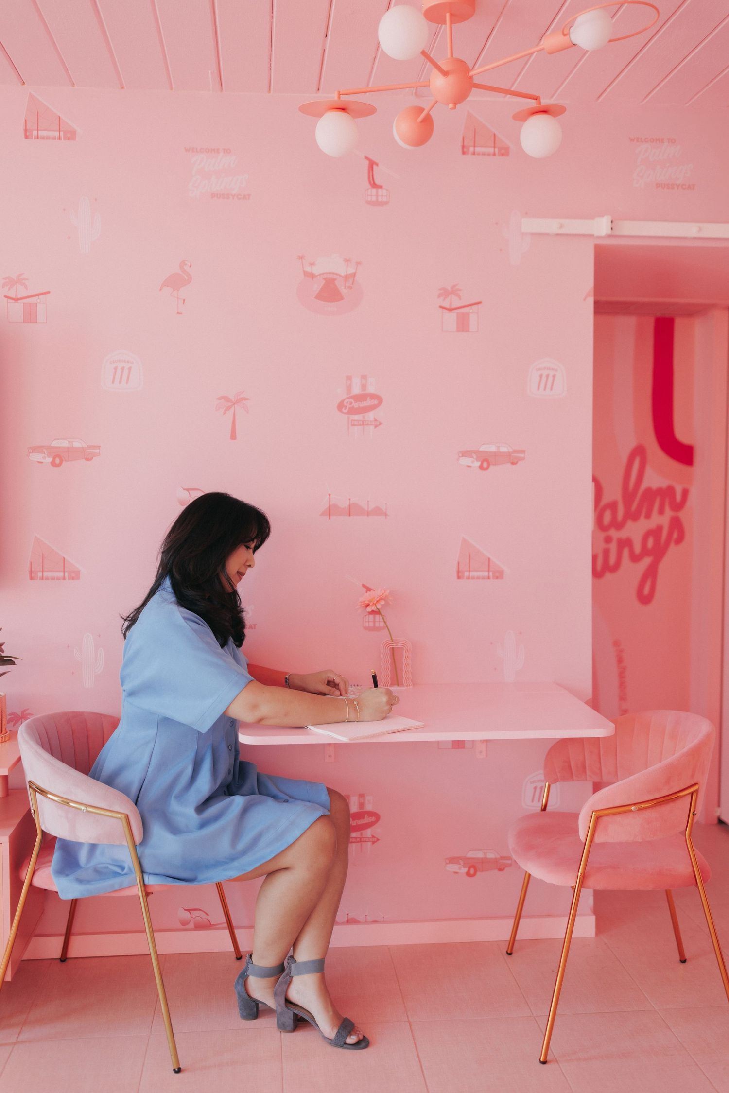 Woman in blue dress writing at a pink table in a pink retro-themed cafe with flamingo wallpaper.