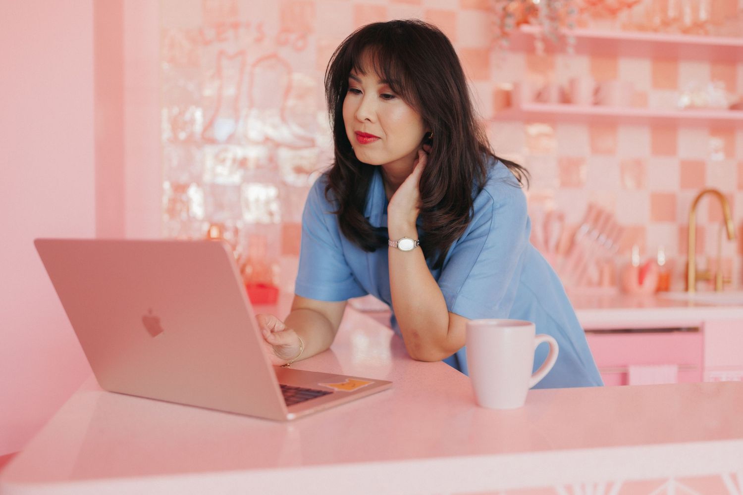 A young Asian woman in a blue top works on a pink MacBook laptop in a pink-themed cafe setting.
