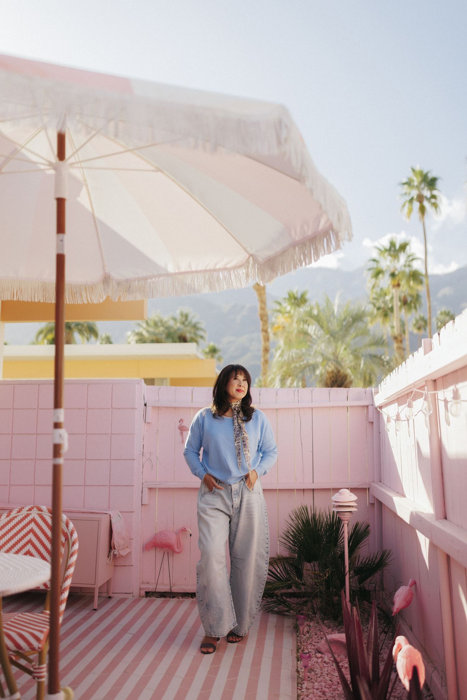 Woman in blue shirt and wide-leg jeans stands on pink tiled patio with striped umbrella, palm trees, and mountains.
