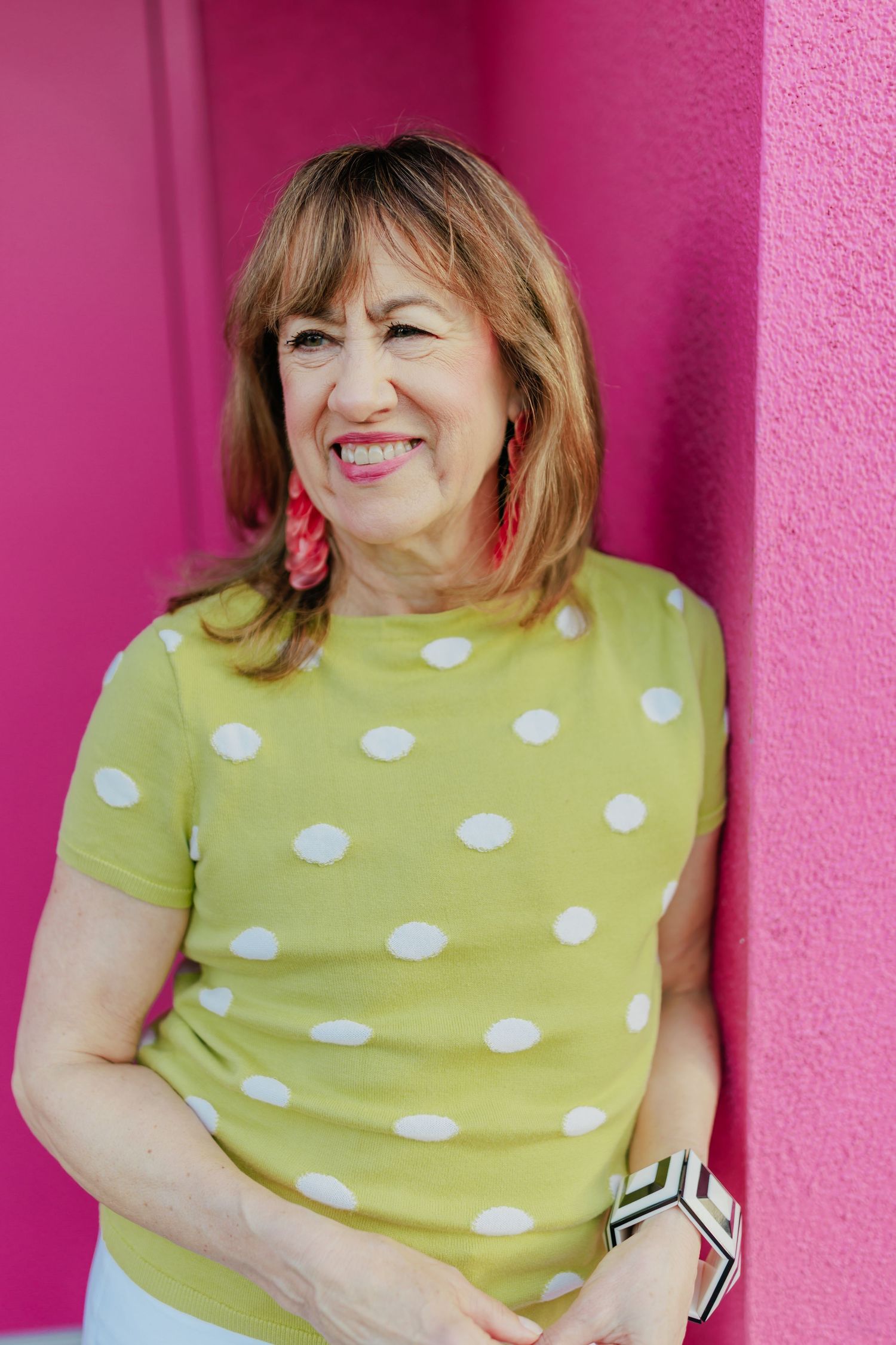Smiling middle-aged woman in green polka dot top and red earrings leaning against a bright pink wall.