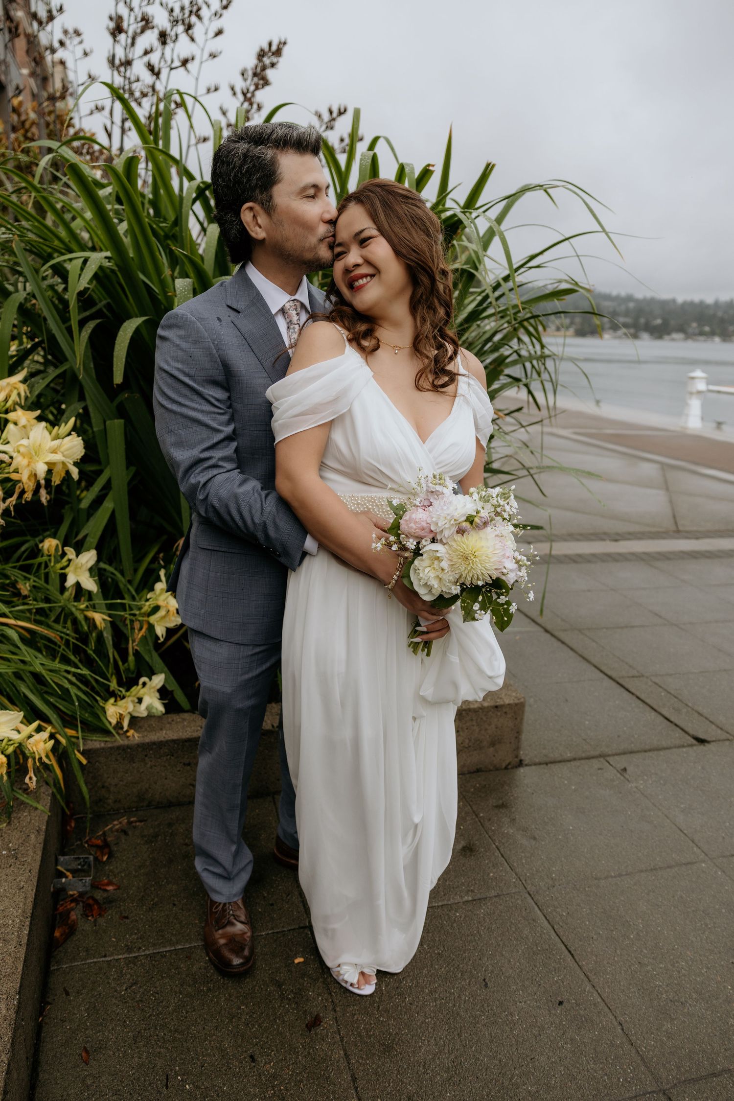 Groom in a gray suit kisses his smiling bride in a white off-the-shoulder gown as she holds a pastel bouquet by the waterfront near Seattle