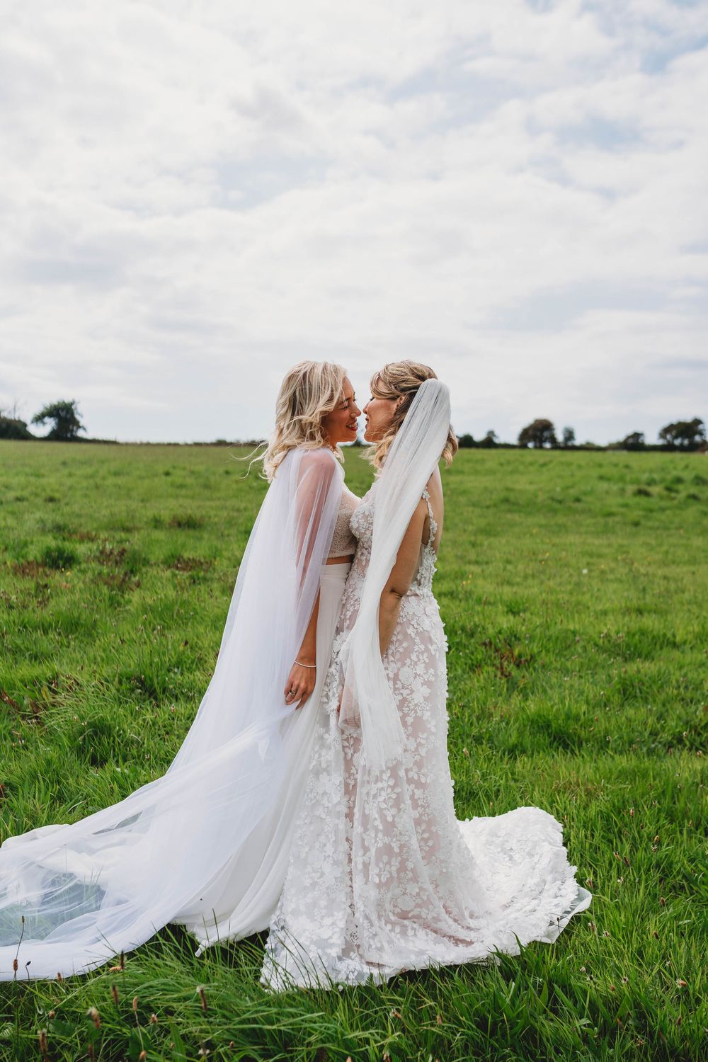 Two brides in lace wedding gowns and flowing veils stand forehead to forehead in a green countryside field under a cloudy sky.