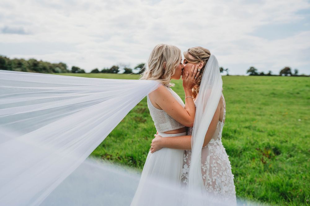 Two brides in white wedding dresses embrace in a green field as a long flowing veil billows in the breeze under a soft cloudy sky.