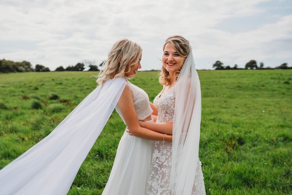 Two brides in white wedding gowns embrace in a green grassy field as a long veil flows in the breeze under a cloudy sky.
