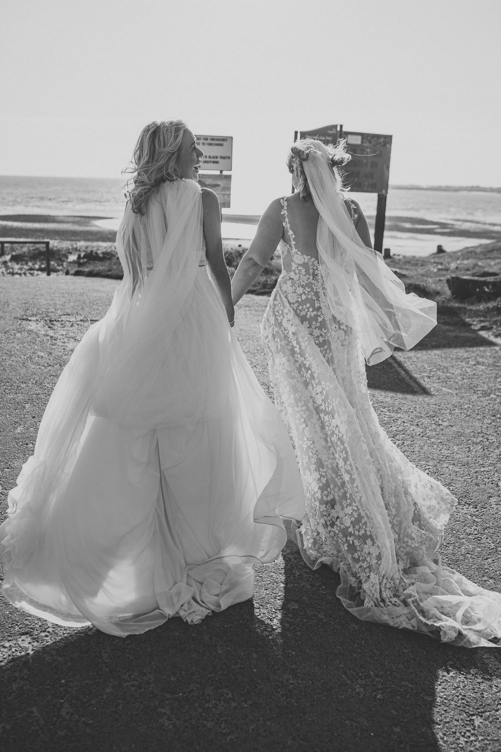 Two brides in flowing lace and tulle wedding gowns with veils walk hand in hand toward the sea in a black and white beach wedding photo.