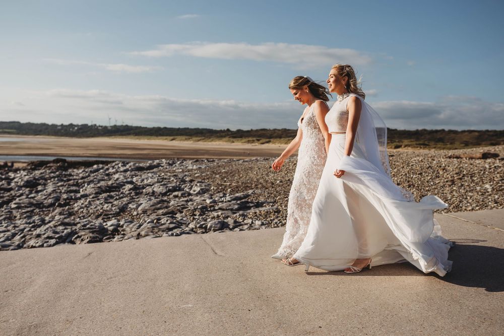 Two brides in white wedding gowns walk hand in hand along a sunlit stone path beside a rocky beach under a soft blue sky.