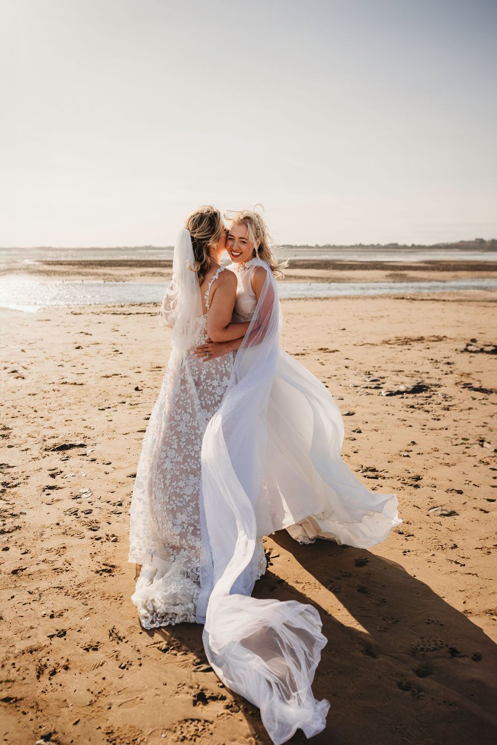 Two brides in flowing lace and tulle wedding gowns embrace and smile on a sunlit sandy beach as the wind lifts their long veils.