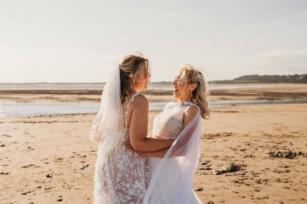 Two brides in white lace wedding gowns and veils embrace and smile on a sunlit sandy beach at golden hour.