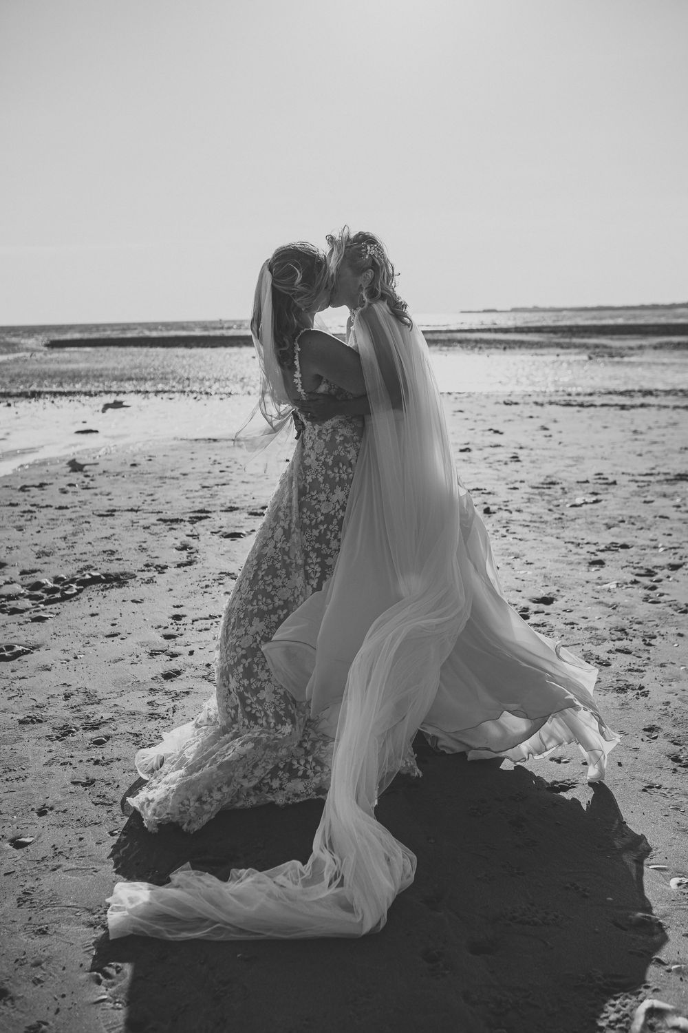 Black and white photo of two brides in lace and tulle wedding gowns embracing on a sunlit beach with flowing veils.