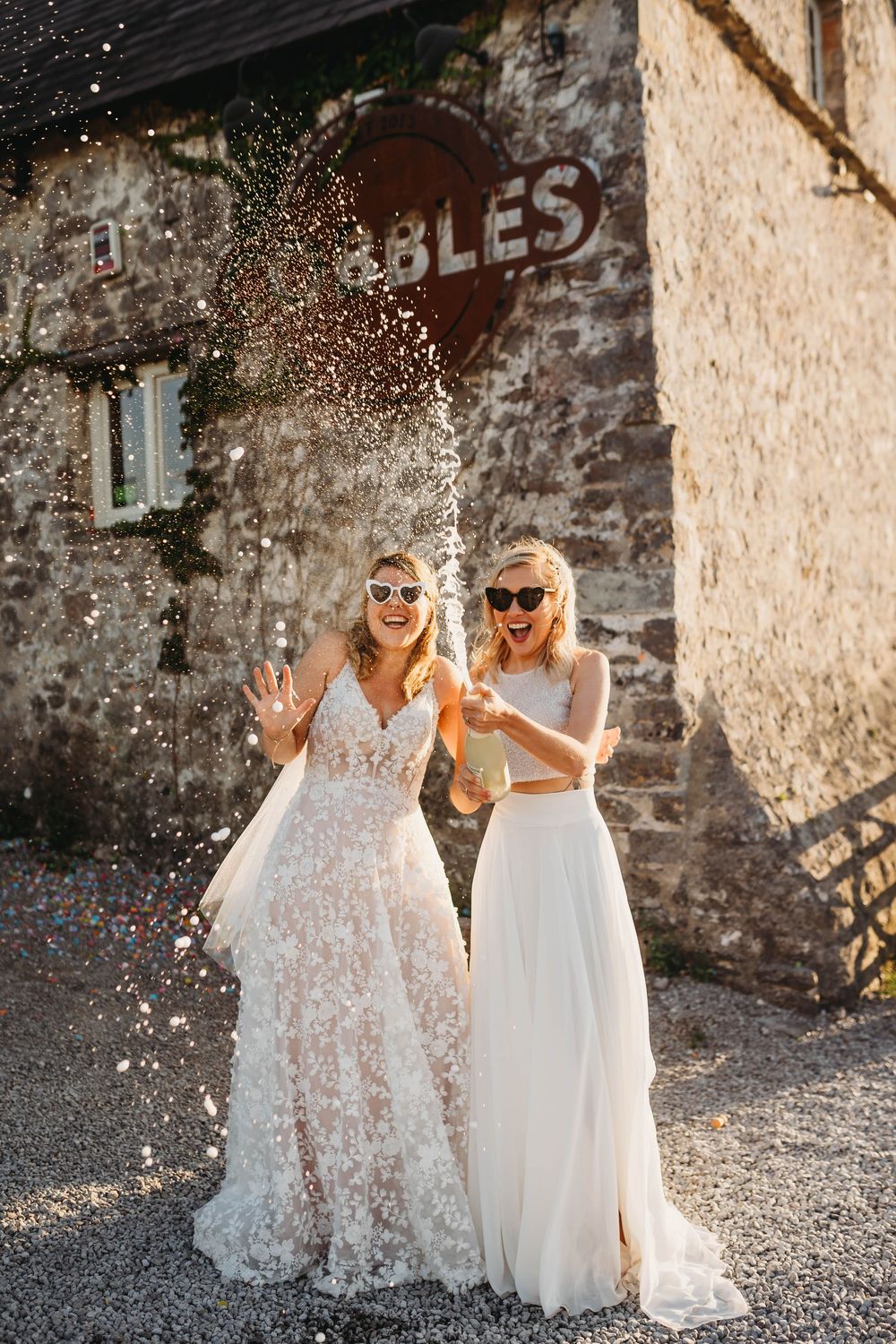 Two brides in lace and chiffon wedding gowns and sunglasses celebrate with a champagne spray outside a rustic stone building.