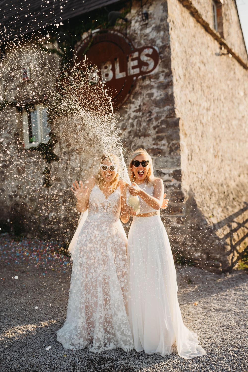 Two brides in lace wedding gowns and sunglasses laugh as they spray champagne in front of a rustic stone building in golden sunlight.