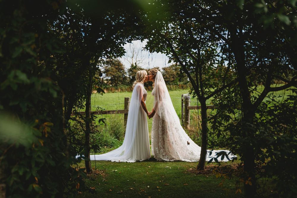 Two brides in white wedding gowns hold hands and face each other beneath leafy trees in a green garden at an outdoor wedding.