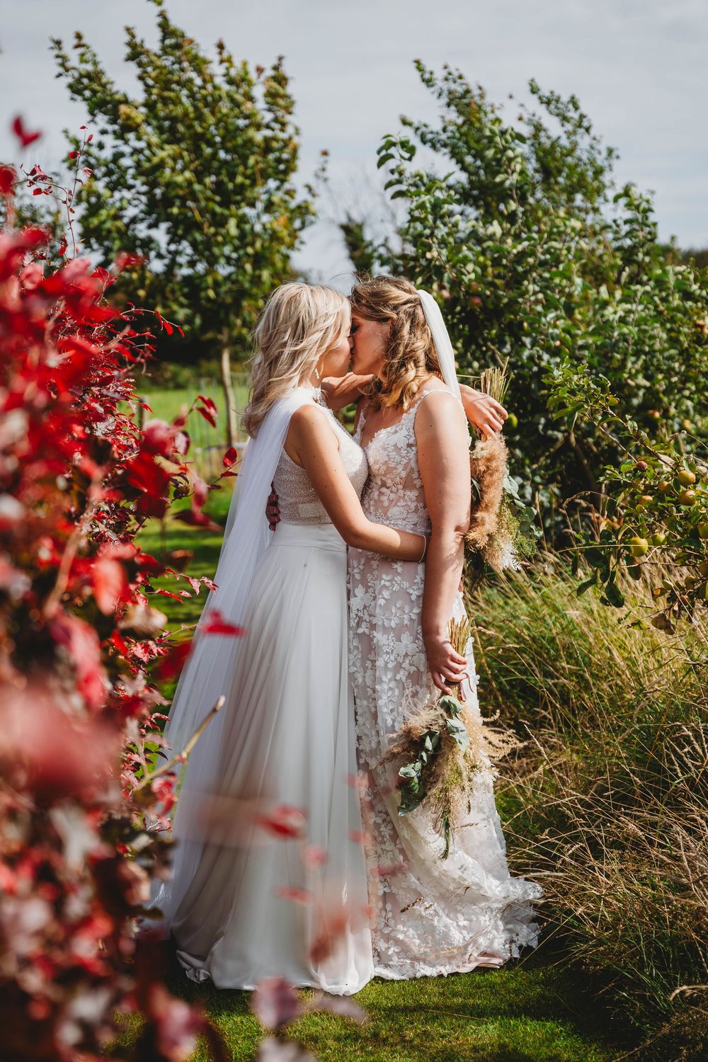 Two brides in wedding dresses and veils share a kiss in a garden beside vibrant red autumn foliage, holding a bouquet.