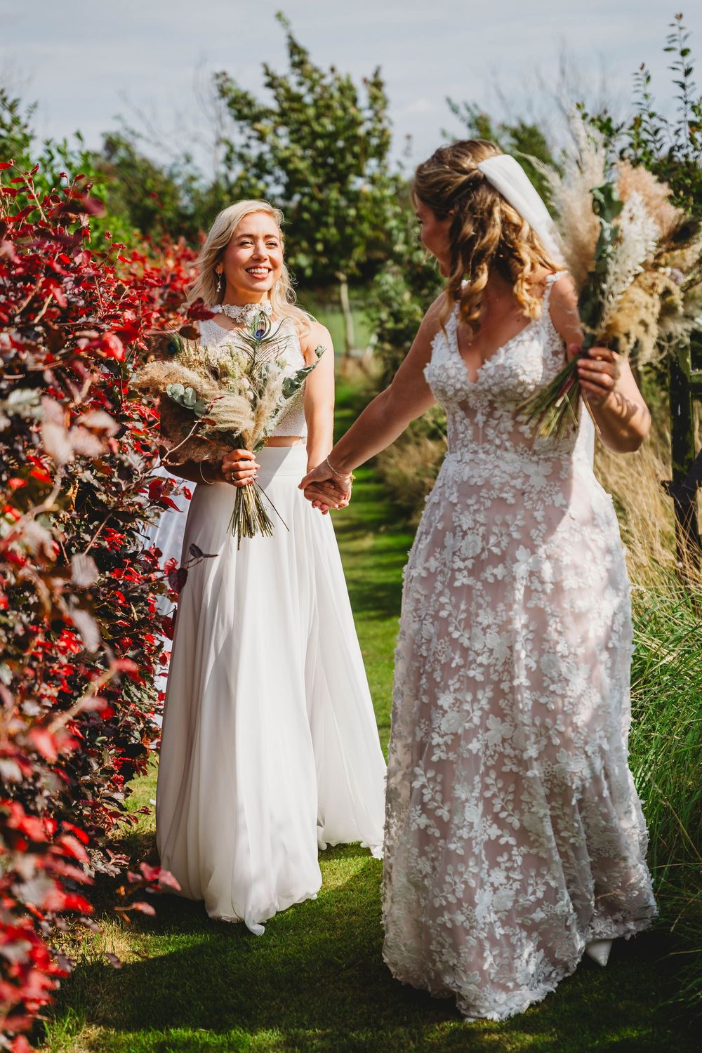 Two brides in floral lace and flowing white wedding dresses hold hands and walk on a grassy path holding pampas grass bouquets.