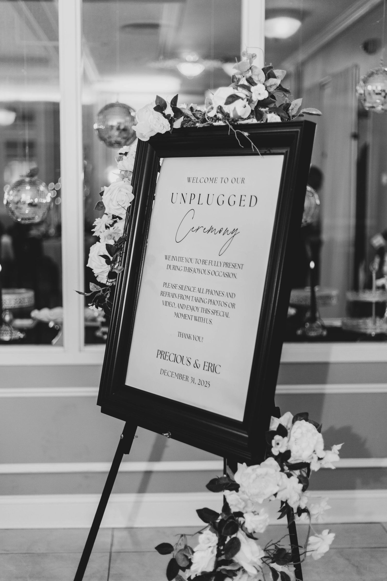 Black and white photo of an unplugged ceremony wedding sign for Precious and Eric, framed on an easel and decorated with white roses.